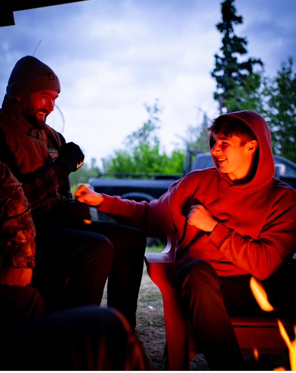 Two generations side by side at the fire, faces warm in the light