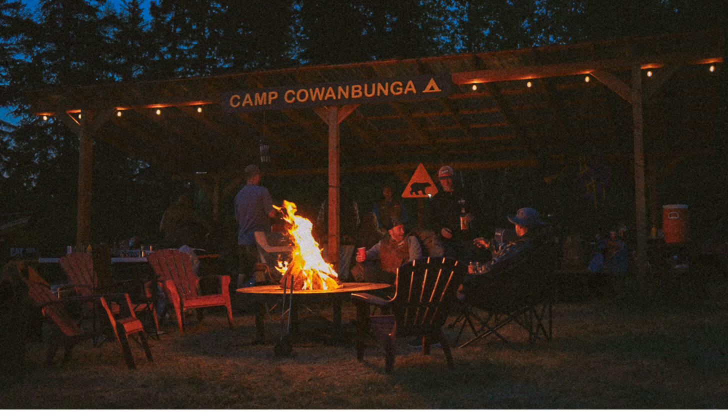 The camp gazebo at night, lit by string lights and a fire, beneath the Camp Cowanbunga sign